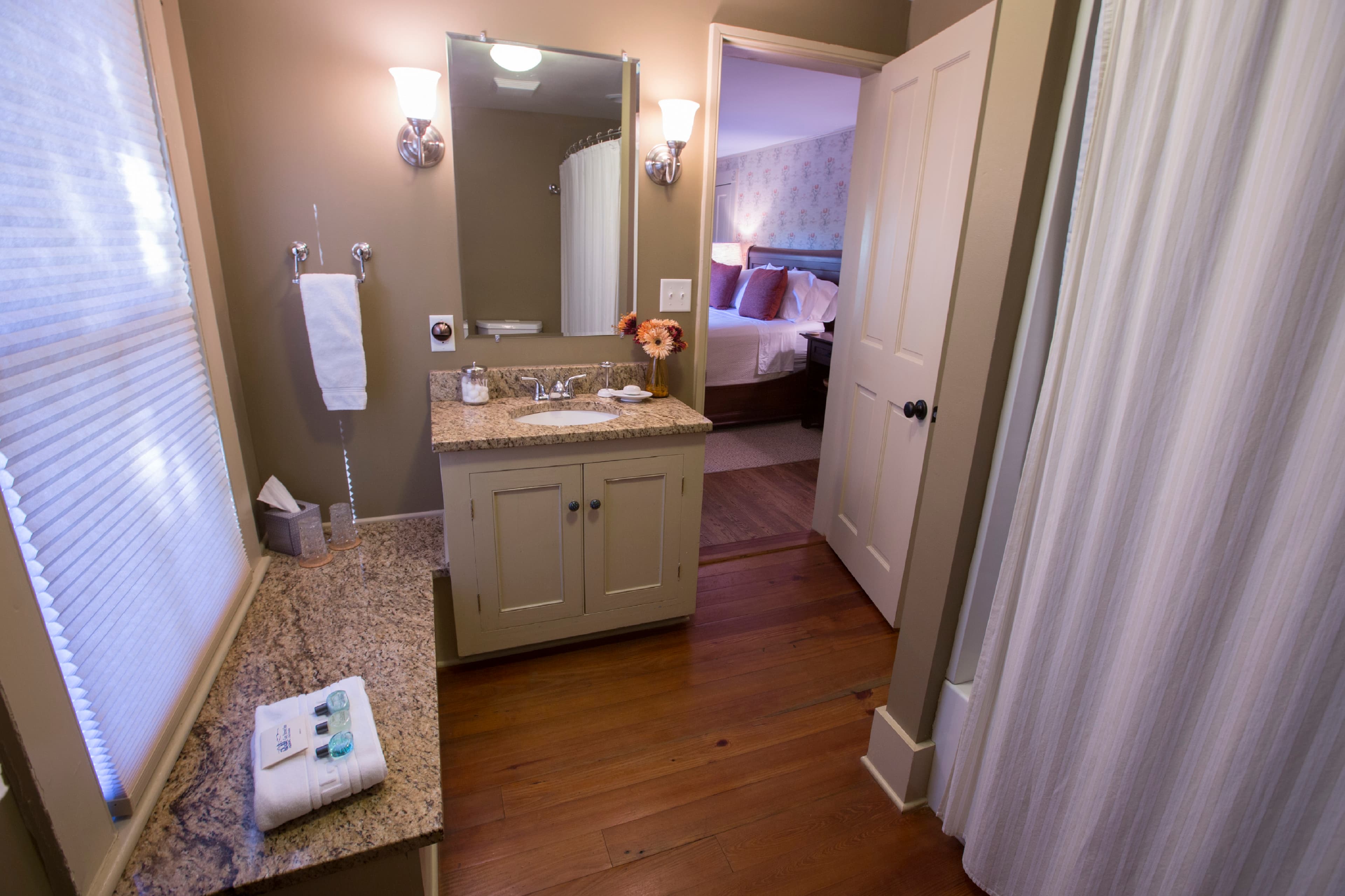 A modern bathroom featuring a granite countertop, mirror, and a view of a bedroom through an open door.