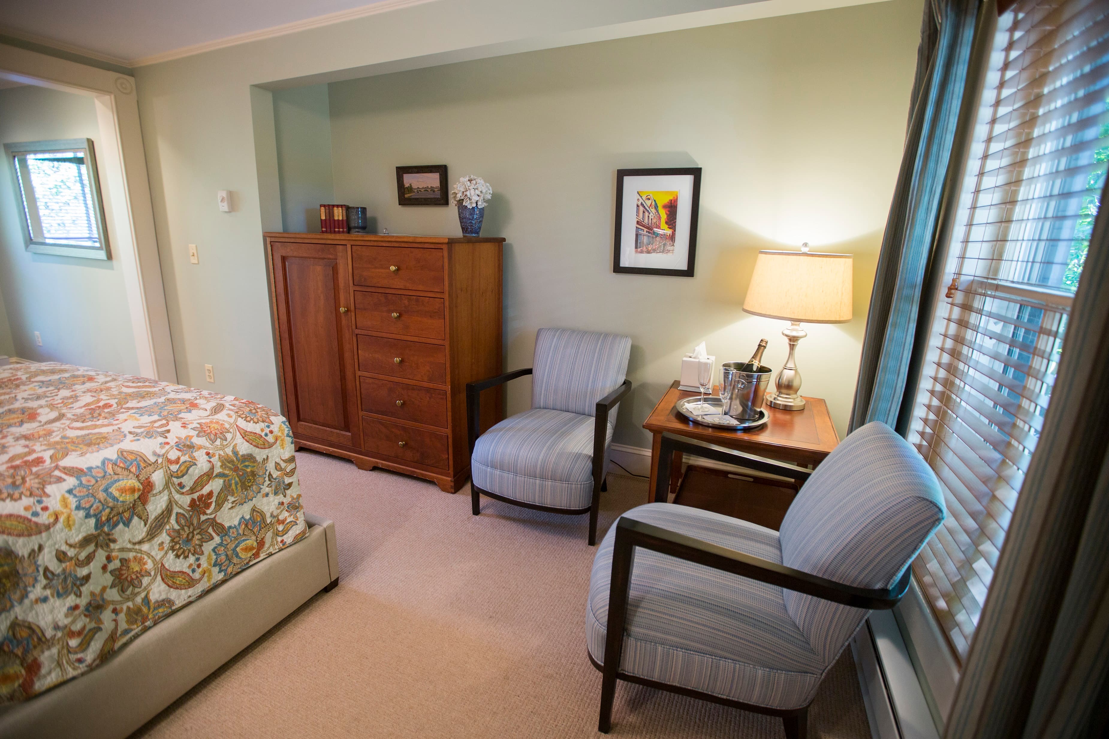 A cozy bedroom featuring a patterned bedspread, a wooden dresser, two armchairs, and a bedside table with a lamp.