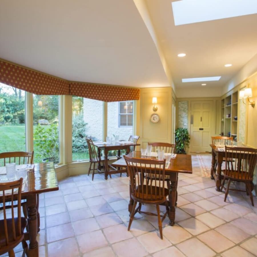 A cozy dining area with wooden tables, chairs, and large windows overlooking a garden.