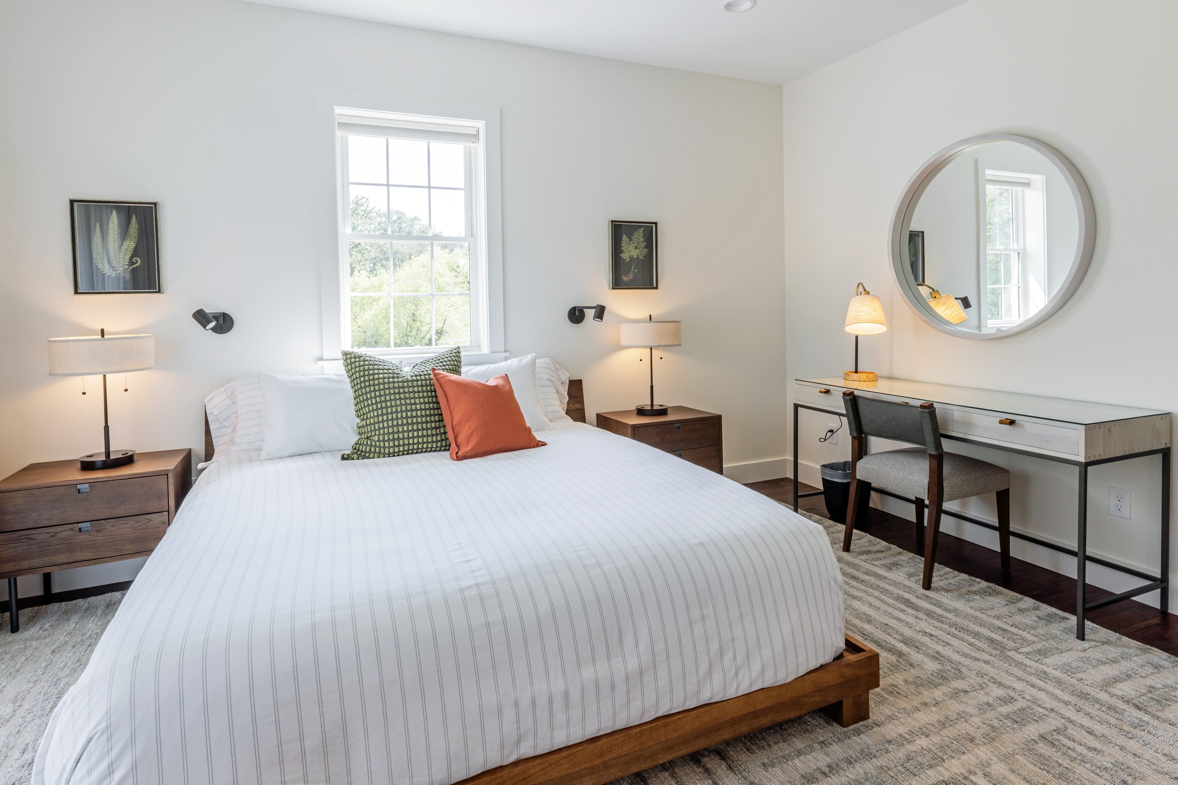 A bright, modern bedroom featuring a queen-sized bed, bedside tables with lamps, and a small desk with a mirror.