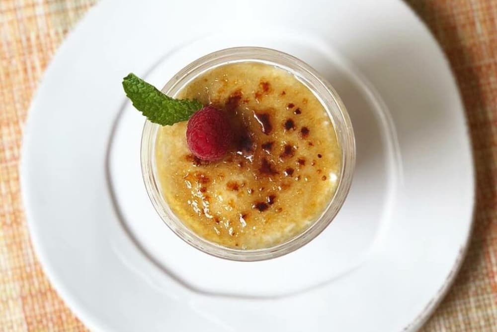 A creamy dessert topped with a caramelized layer, garnished with a raspberry and mint leaf.