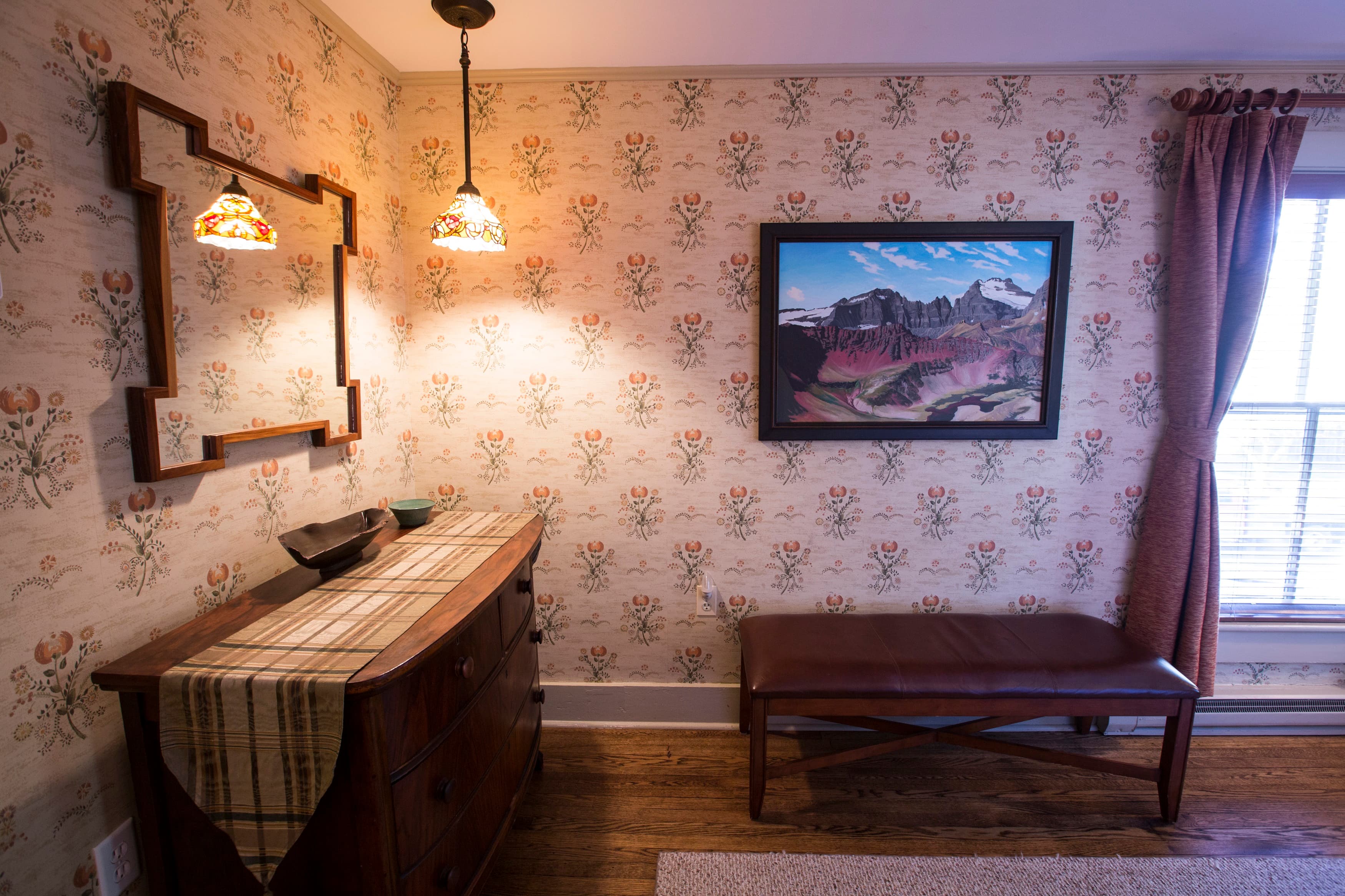 Cozy room featuring floral wallpaper, a wooden dresser, a bench, and a landscape painting.