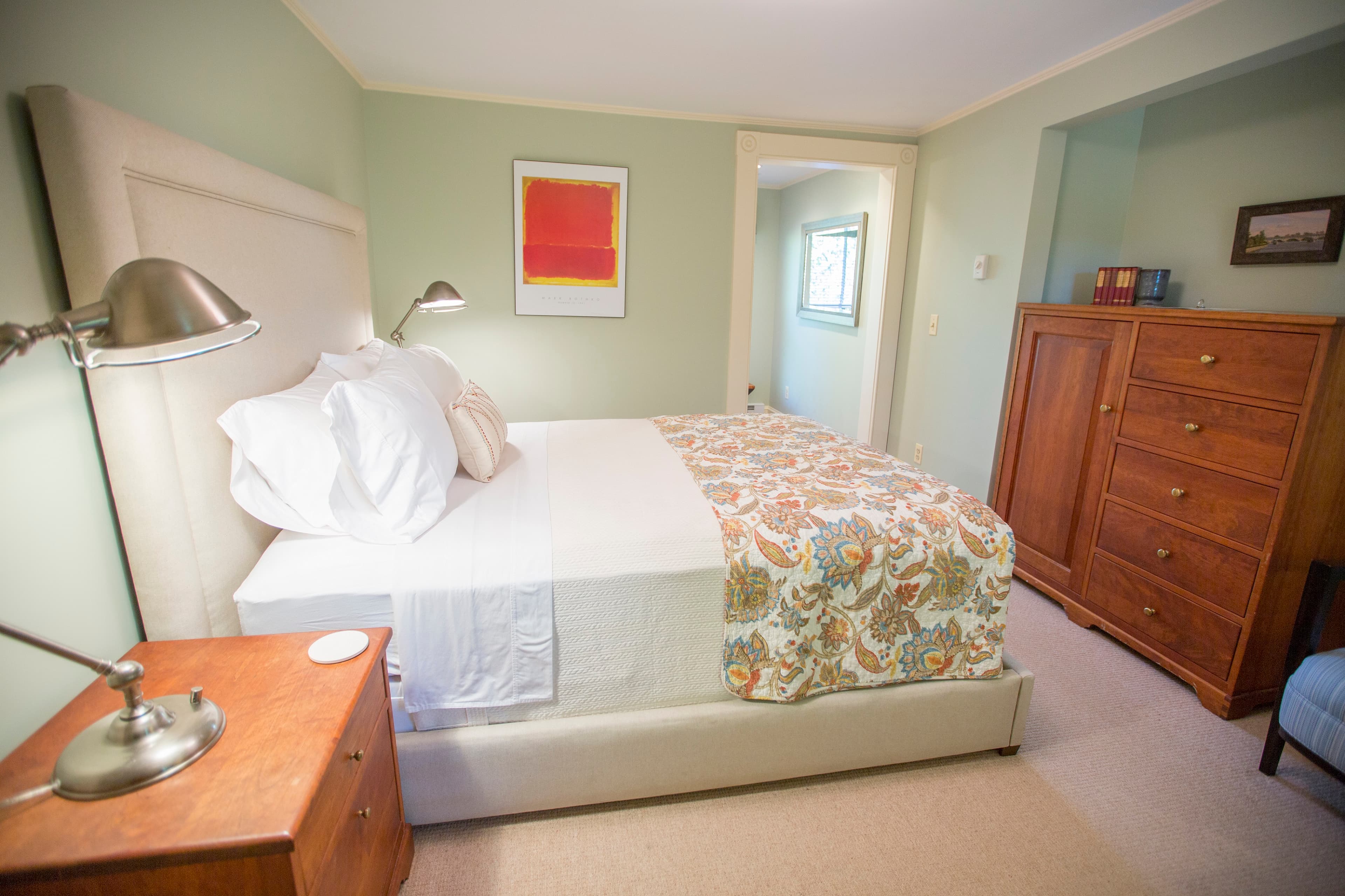 A neatly arranged bedroom featuring a bed with a colorful blanket, a bedside table with a lamp, and a wooden dresser against a light green wall.