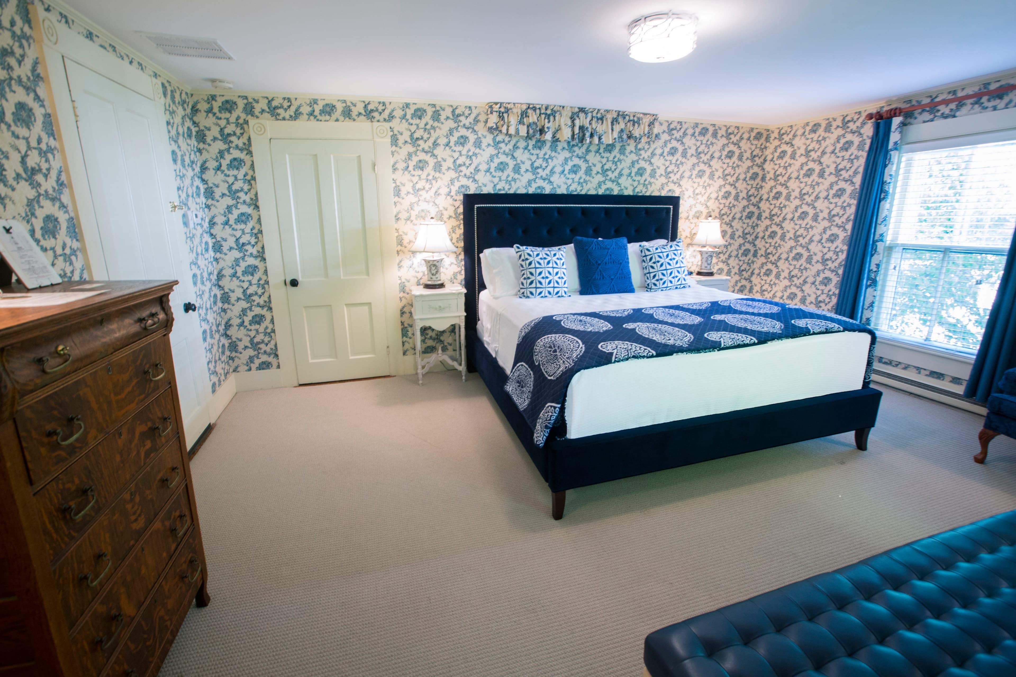 A cozy bedroom featuring floral wallpaper, a navy upholstered bed, and vintage furniture.