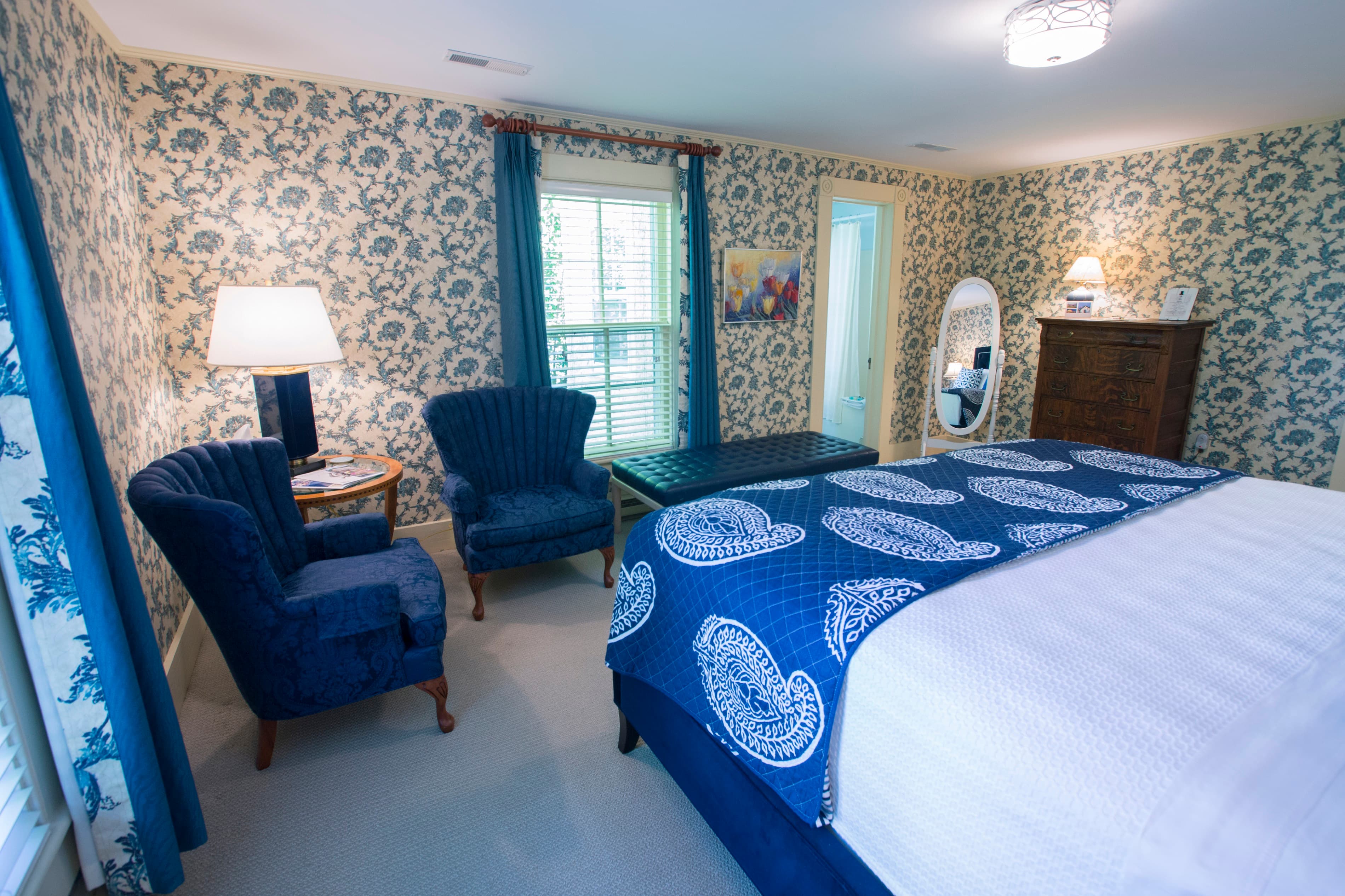 A cozy bedroom featuring floral wallpaper, a blue bedspread, two blue armchairs, and a wooden dresser.