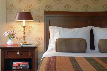 A cozy bedroom with a wooden headboard, decorative pillows, a nightstand, a lamp, and a bouquet of flowers.