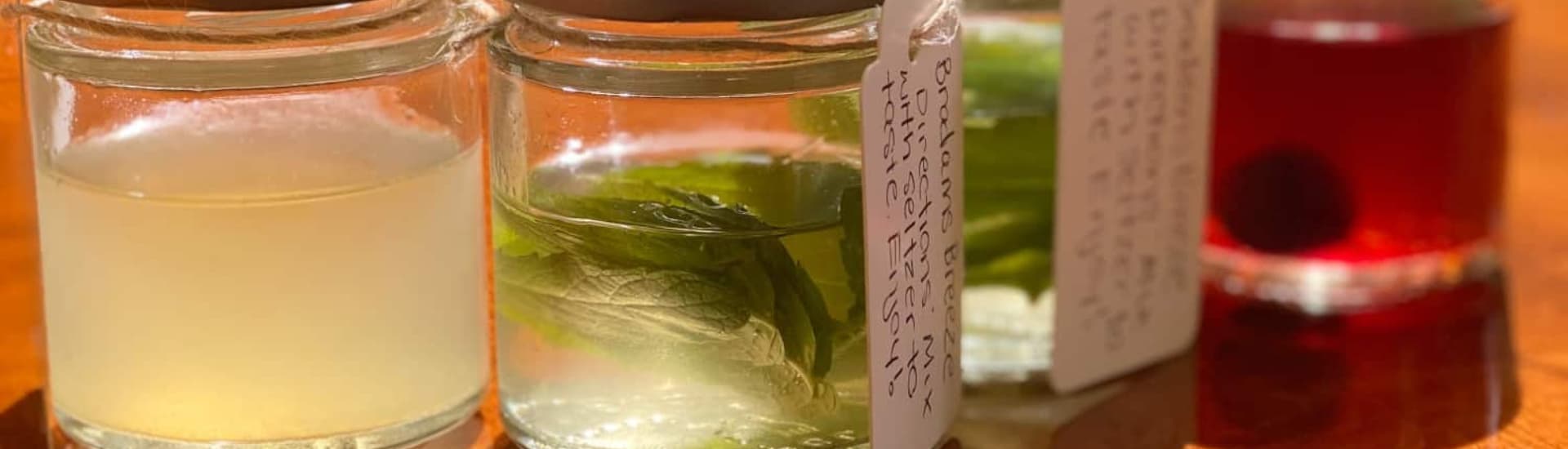 Three small glass jars containing flavored liquid infusions, with a red drink on the right and mint leaves visible in the center jar.