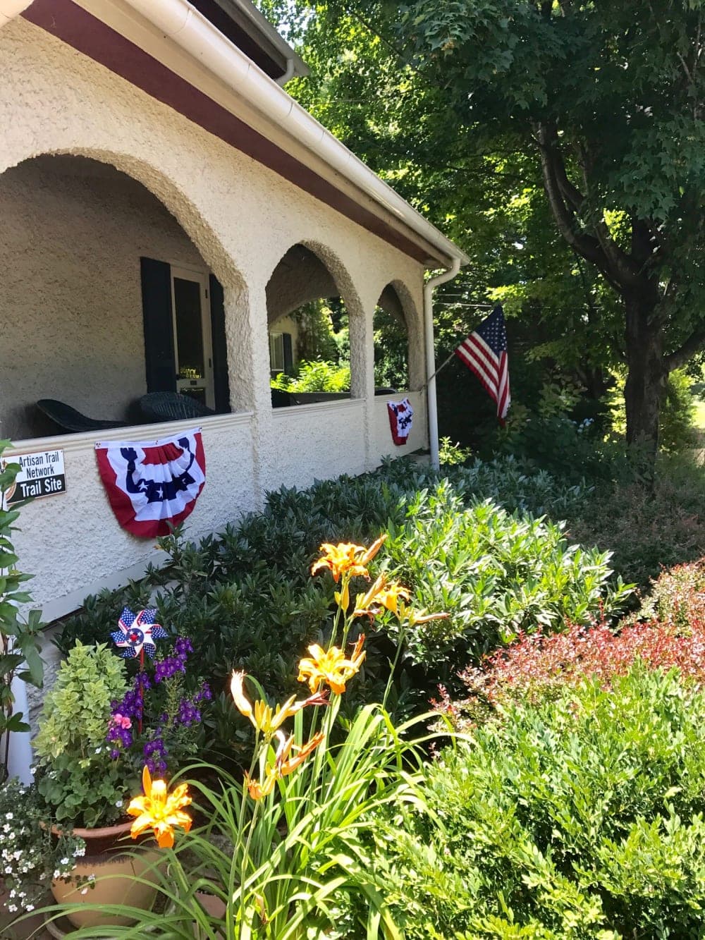 A porch adorned with patriotic bunting and an American flag, surrounded by colorful flowers and greenery.