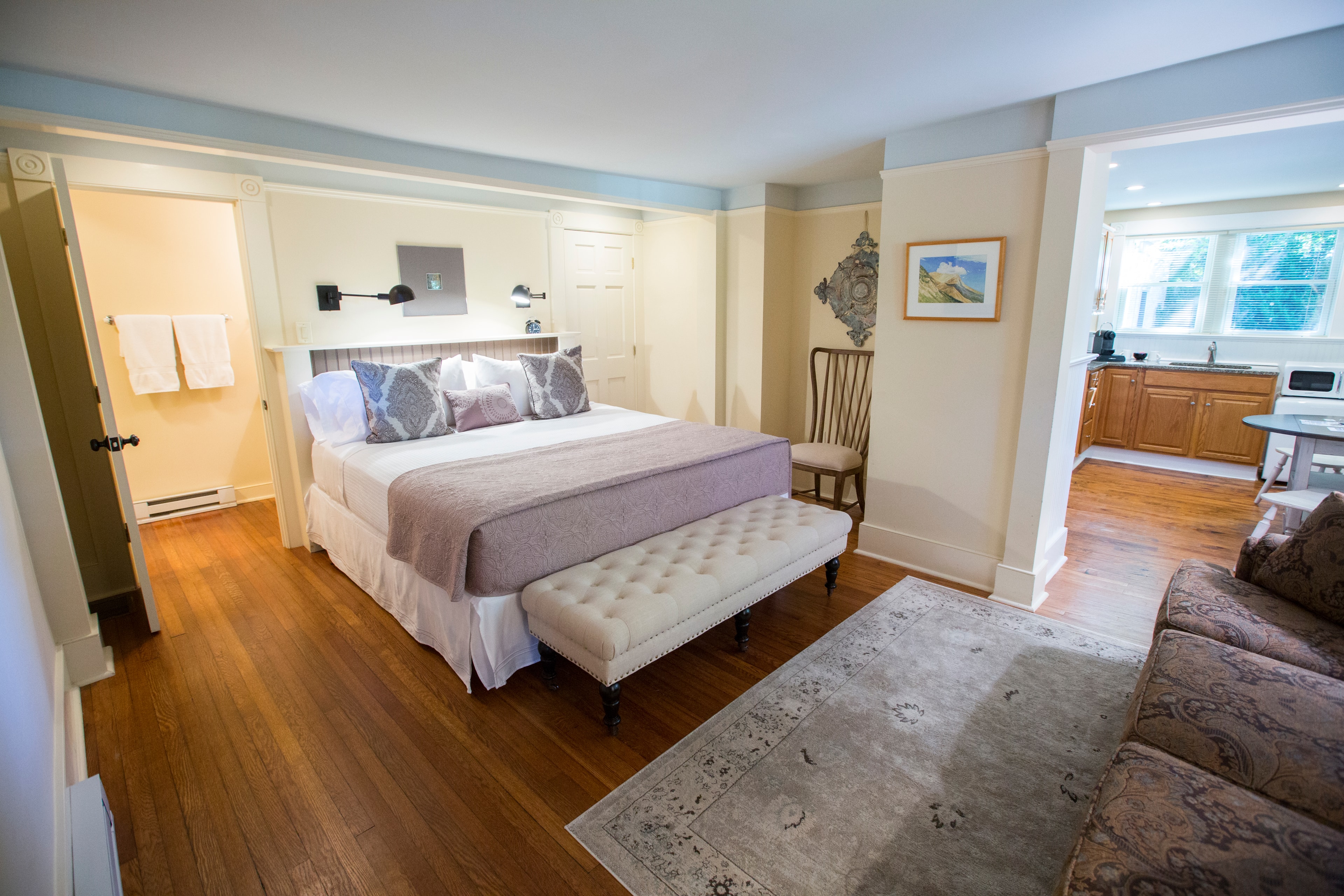 Bright and cozy bedroom with a king-size bed, a bench, and a view into a kitchen area.