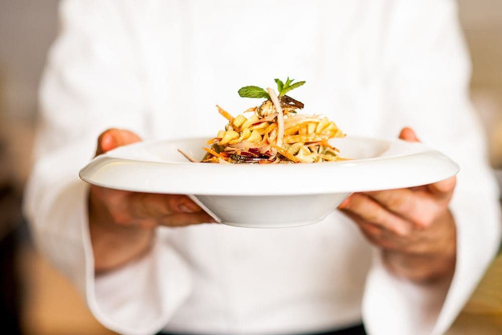 A chef presents a dish of pasta garnished with herbs on a white plate.