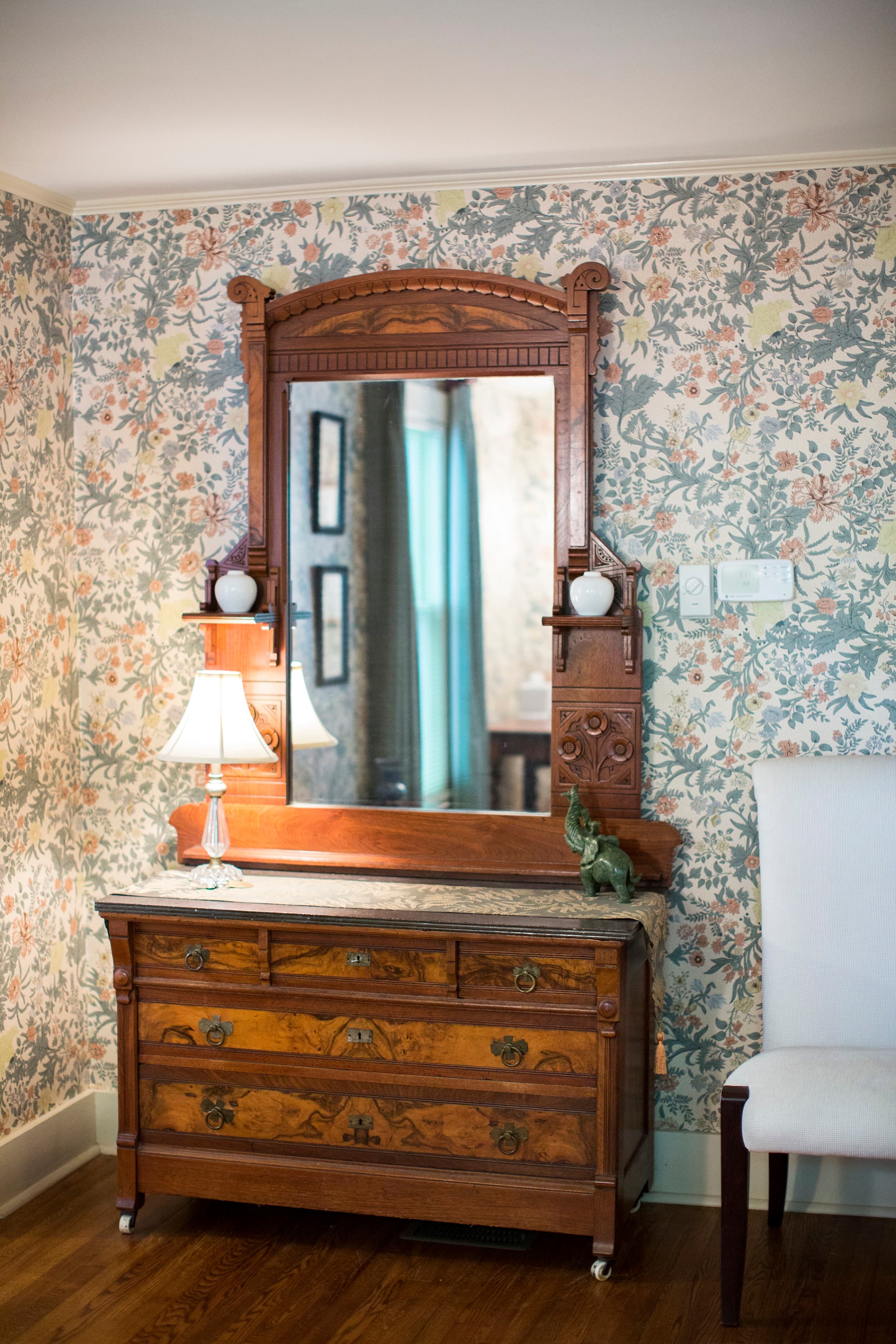 An ornate wooden dresser with a mirror and lamp against a floral wallpaper backdrop.