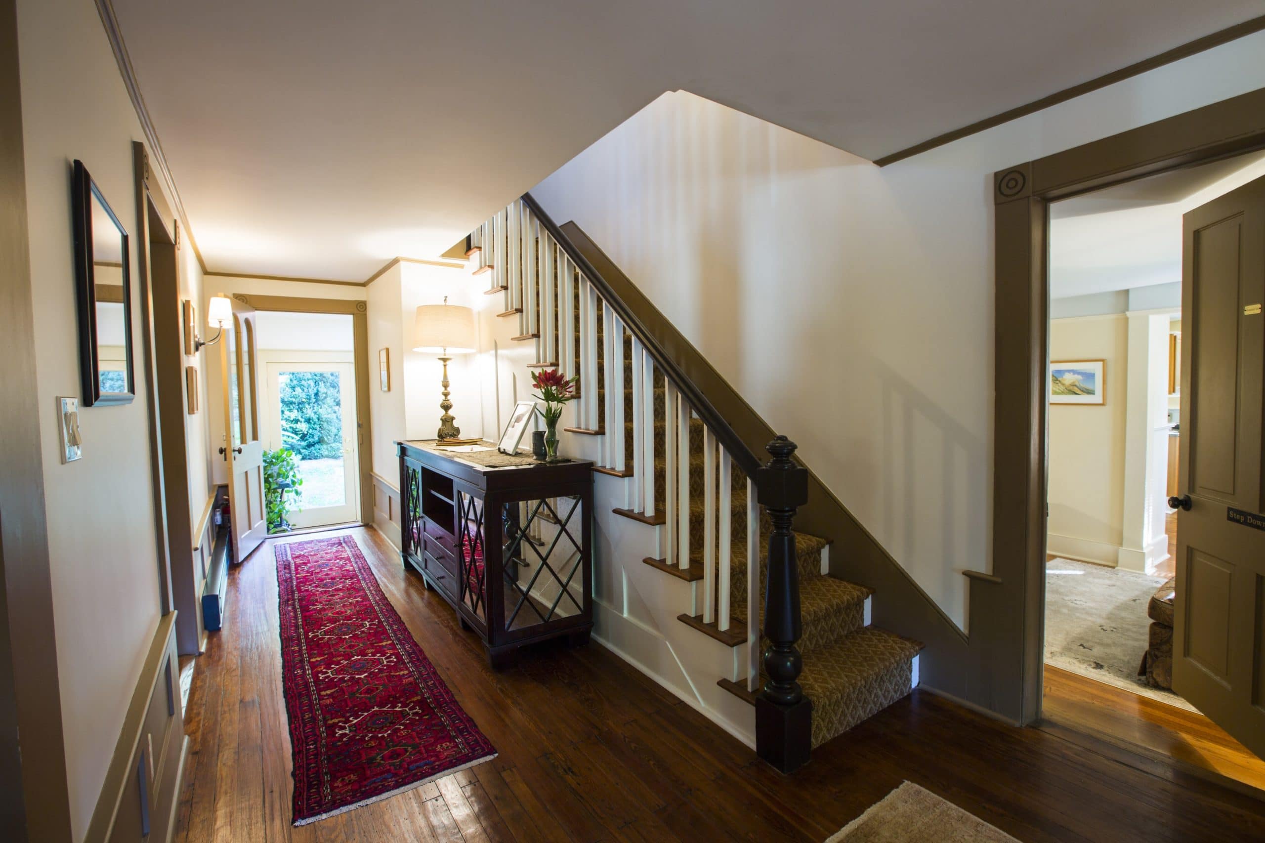 A hallways with a piece of furniture with a lamp and flowers on it, a staircase going up to another floor and an open doorway into another room.