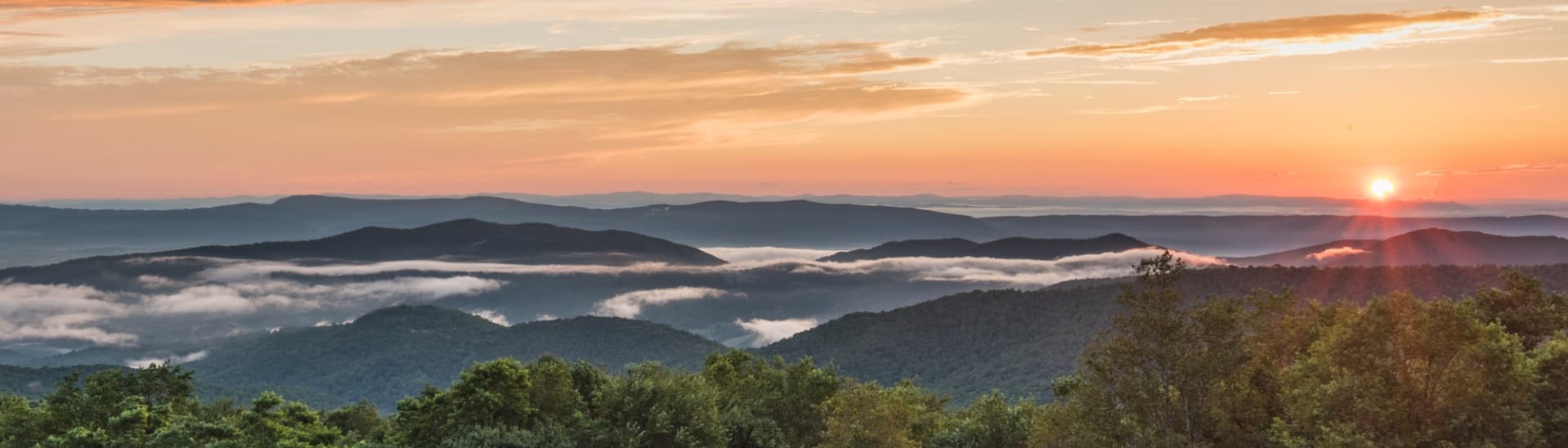 A panoramic view of rolling mountains and valleys under a colorful sunrise with mist in the lowlands.