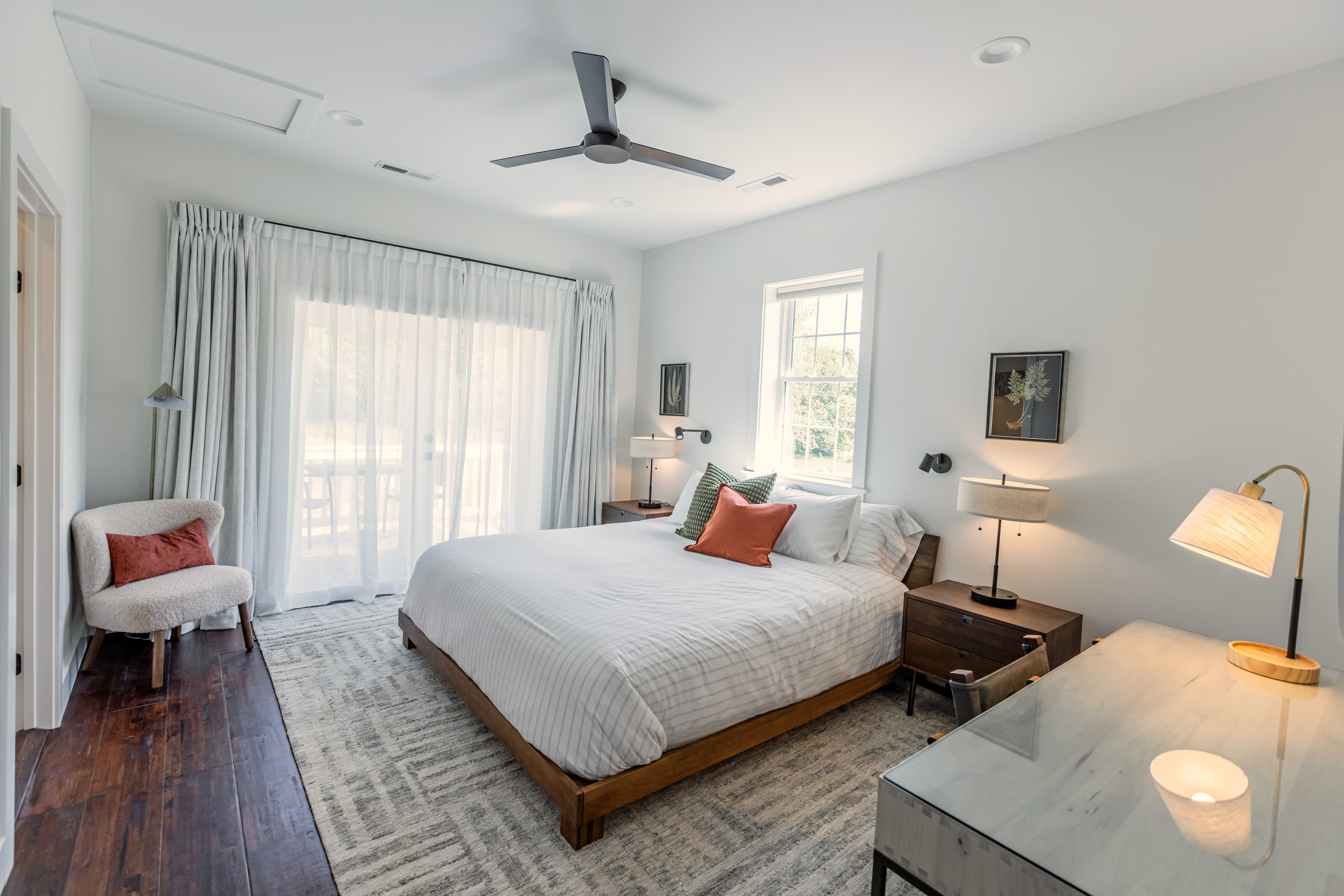 A modern, airy bedroom featuring a large bed, bedside lamps, and a stylish desk with a chair, illuminated by natural light from windows.