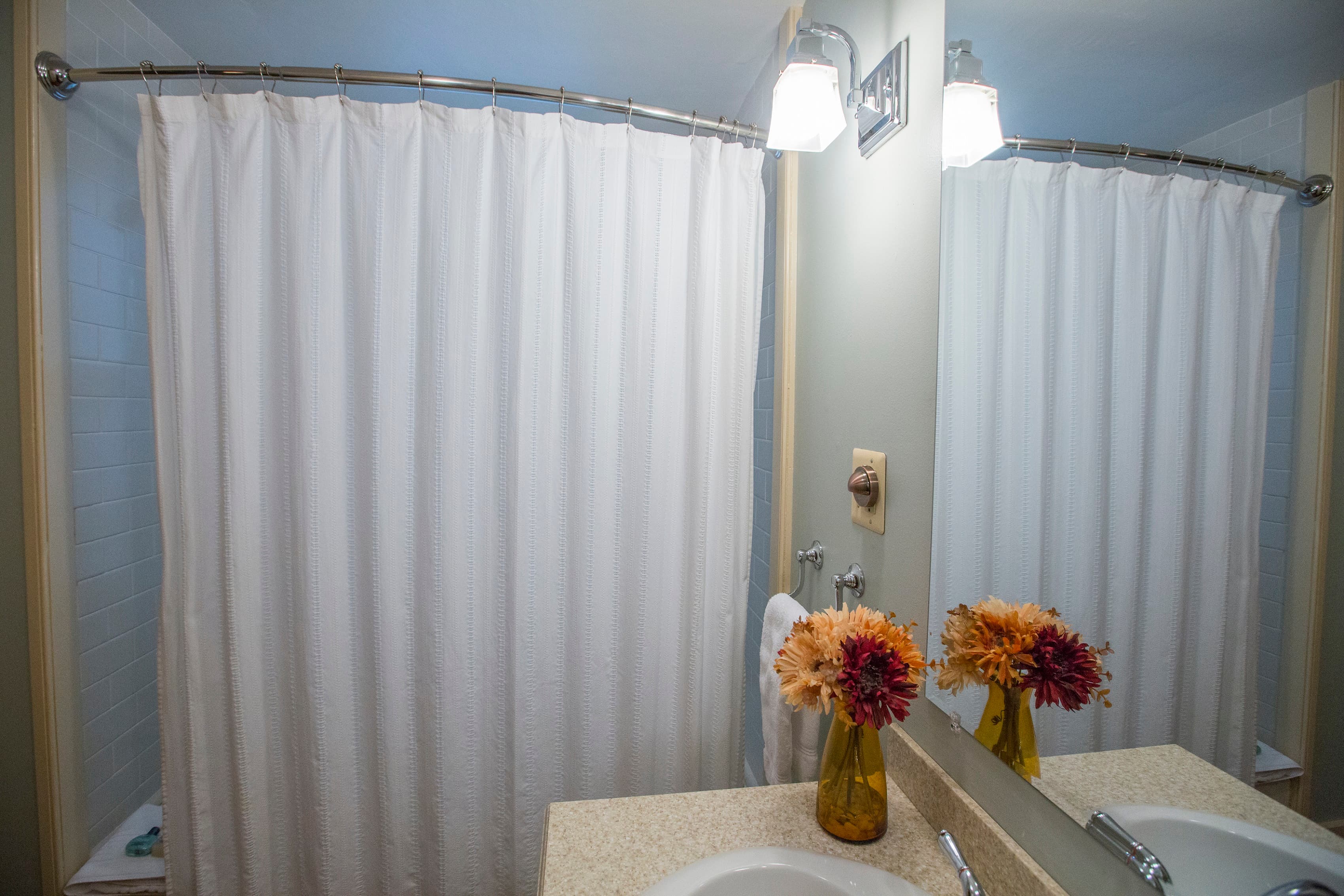 A bathroom with a white shower curtain, a sink with a mirror, and a vase of flowers on the countertop.