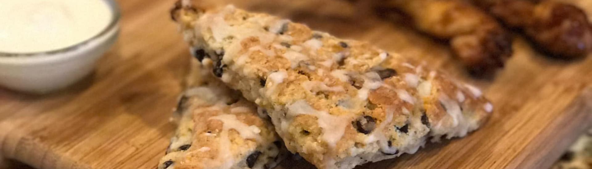 Two scones drizzled with icing on a wooden board next to a small bowl of cream.