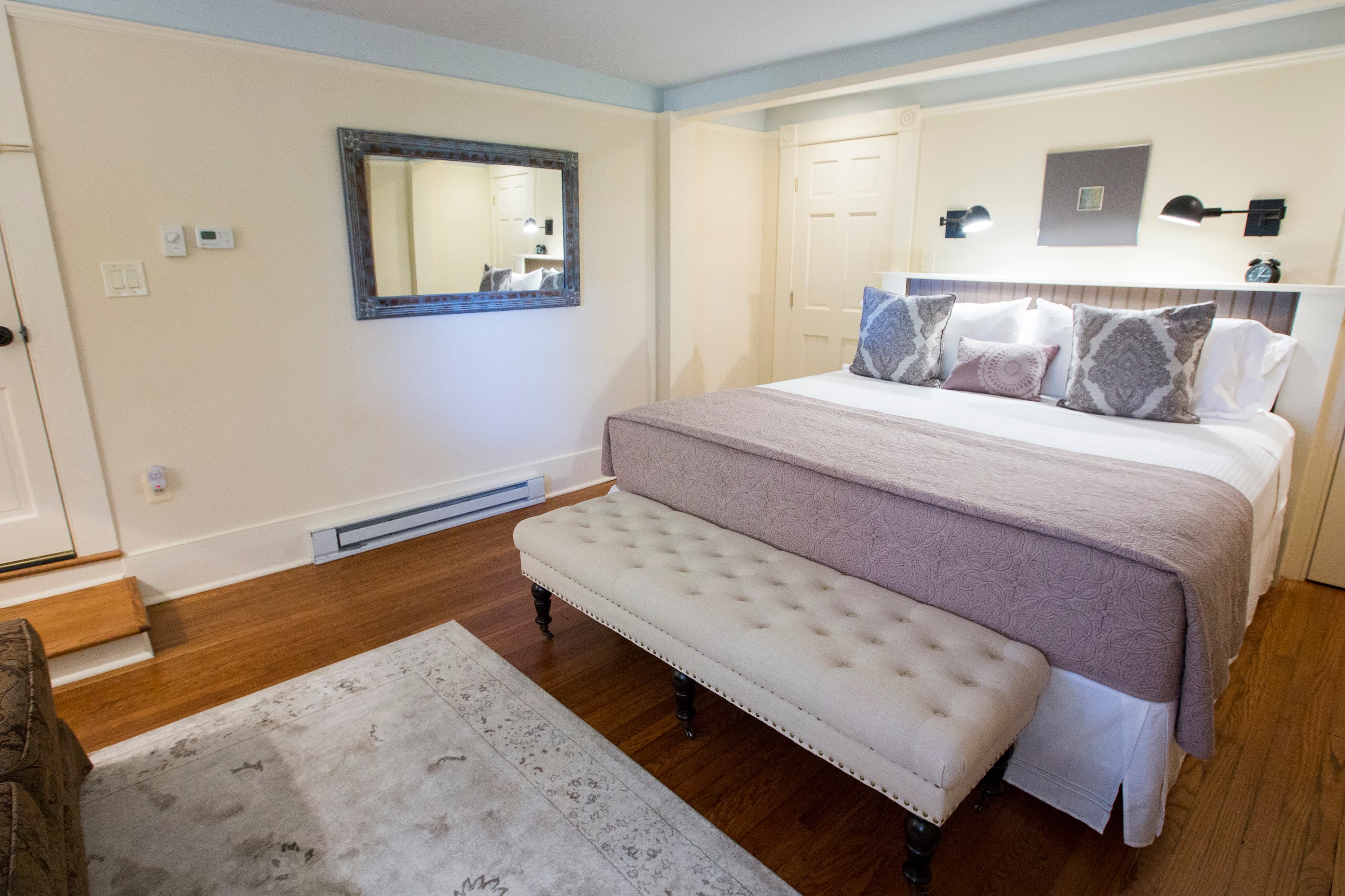 A cozy bedroom featuring a bed with decorative pillows, a bench at the foot, a mirror, and wooden flooring.