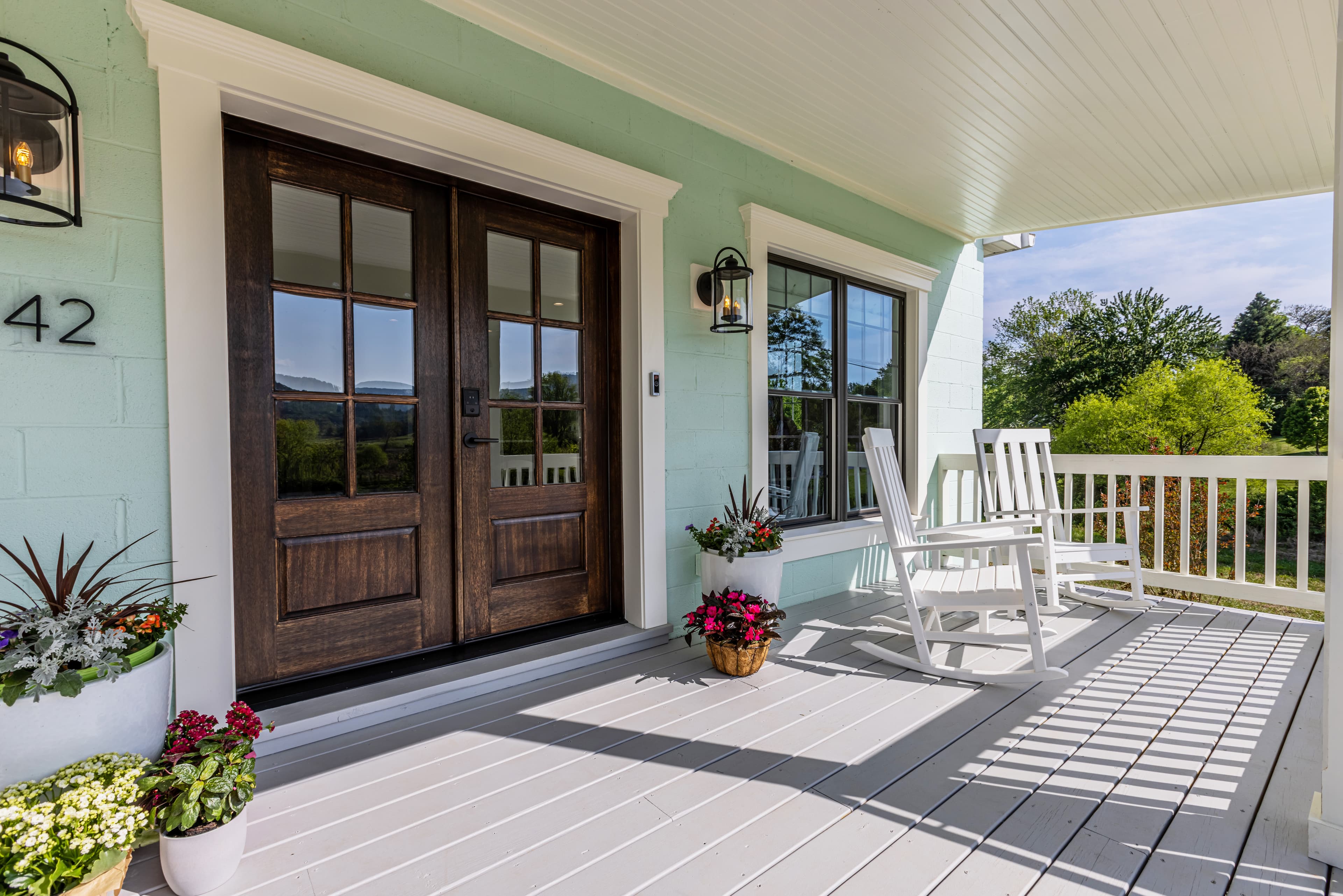 A welcoming porch with double wooden doors, white rocking chairs, and potted flowers against a light green wall.