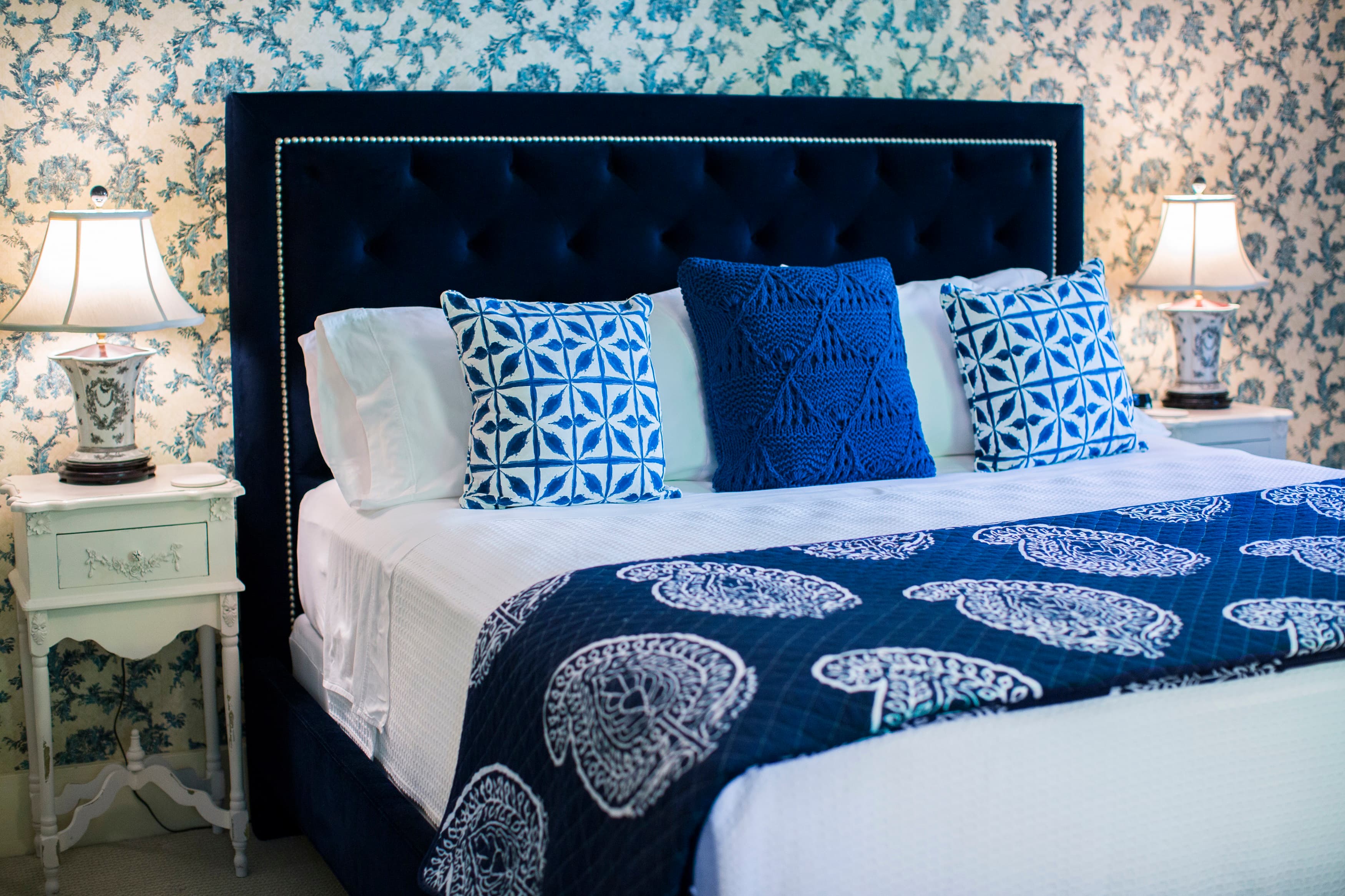 A stylish bedroom featuring a navy tufted headboard, blue and white patterned pillows, and a floral wallpaper backdrop.